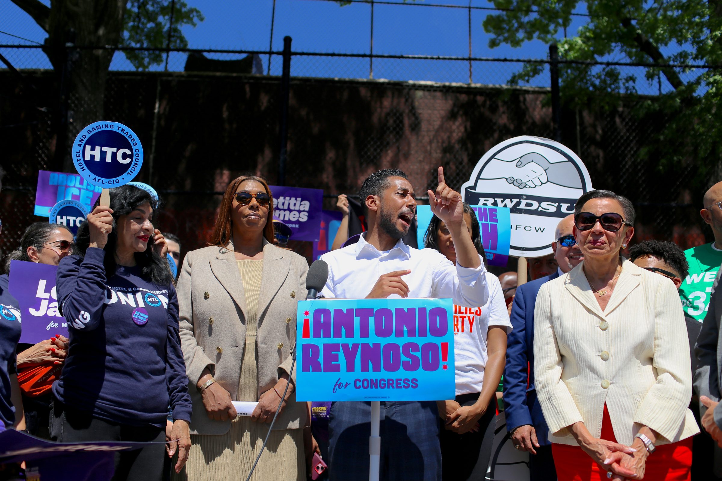 Antonio Reynoso speaking at a rally podium behind an 'Antonio Reynoso for Congress' sign, surrounded by supporters